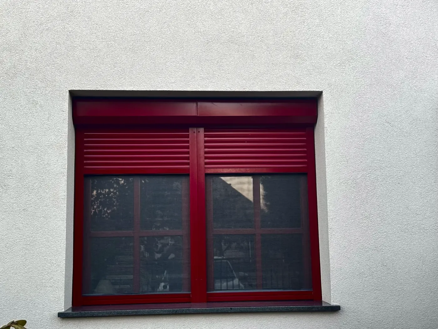 Bordó színű redőny egy bordó keretes ablakon, fehér homlokzaton